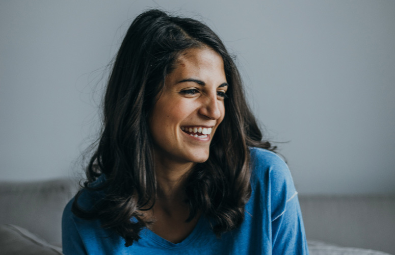 Dark-haired young woman wearing a blue sweater looks to the side while smiling about dental implants