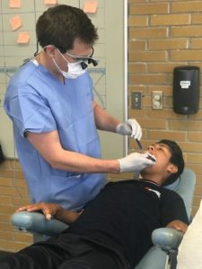 Dr. Moos wears powder blue scrubs as he performs dental work on a young man in a black shirt in Puebla, Mexico
