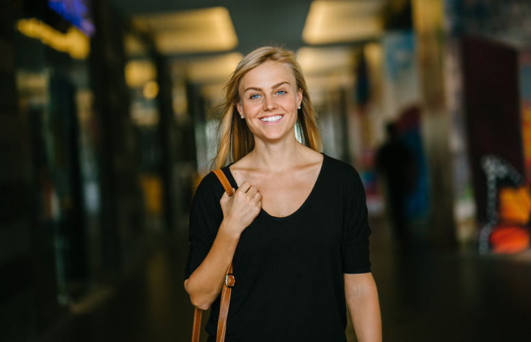 Blonde woman wearing a black shirt and brown shoulder bag smiles with custom-made dental veneers