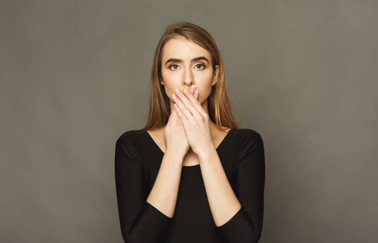 Blonde woman with a loose adult tooth covers her mouth with her hands while wearing a black shirt