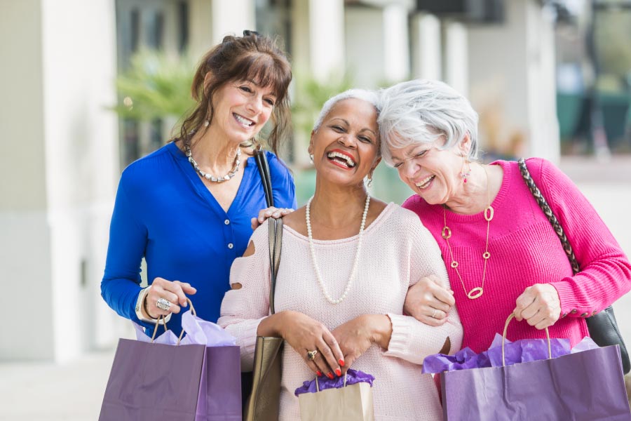 3 Older laughing women of mixed ethnicity walking arm in arm down the sidewalk with shopping bags