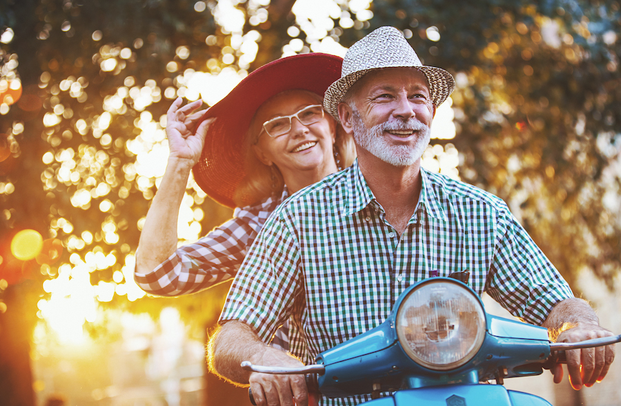 Closeup low angle front view of mid 60's couple cruising a scooter bike on a summer afternoon. They are spending their retirement days enjoying the life. Sunlight is flaring from background.